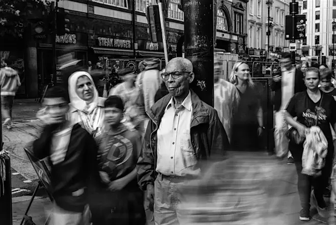 Man is leaning on a street light. Around him walking a lot of people