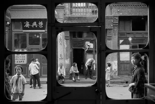 Black-and-white photograph by Marc Riboud showing everyday street life in China, framed through the windows of a building with pedestrians gathered outside.