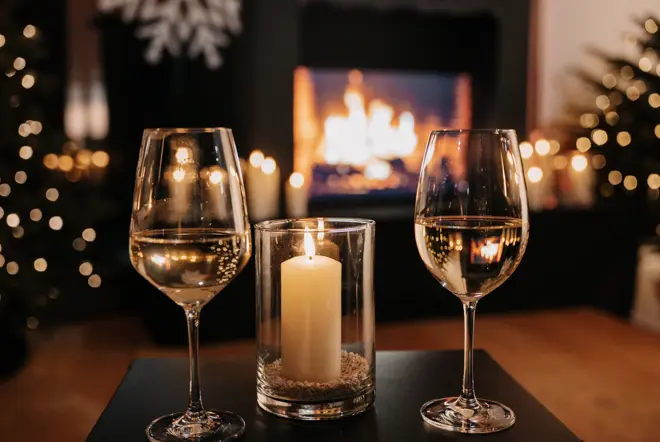 Two filled wine glasses with a candle on the table. A fireplace in the background.