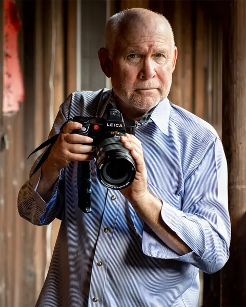 Steve McCurry holds a Leica camera and stands in front of a wooden house.