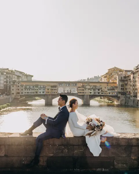 A wedding couple sits on a wall overlooking the river, surrounded by old buildings.