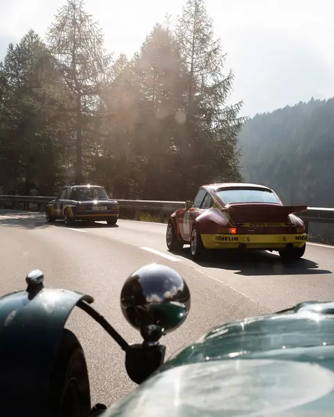 Vintage cars drive along a winding mountain road surrounded by trees and rocks.