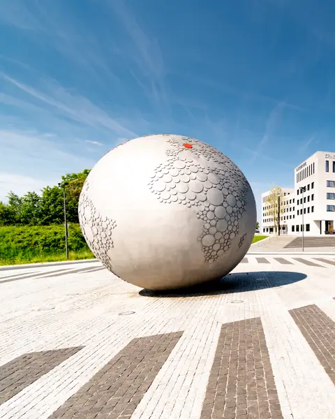 Large stone globe on a square in front of a hotel and parking lot under a blue sky.