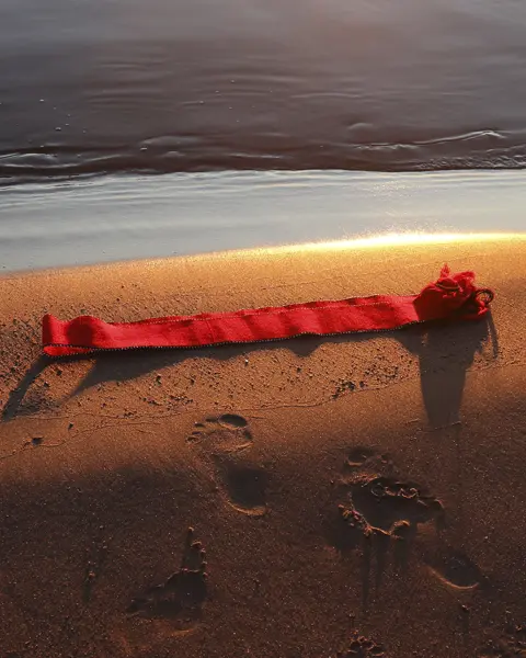 Red ribbon lying on the beach shore at sunset, footprints in the sand.