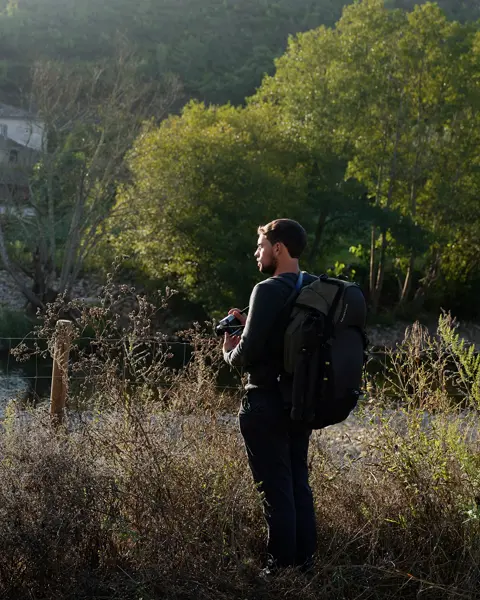 A man with a rucksack stands in the countryside and looks out over a small settlement by the river
