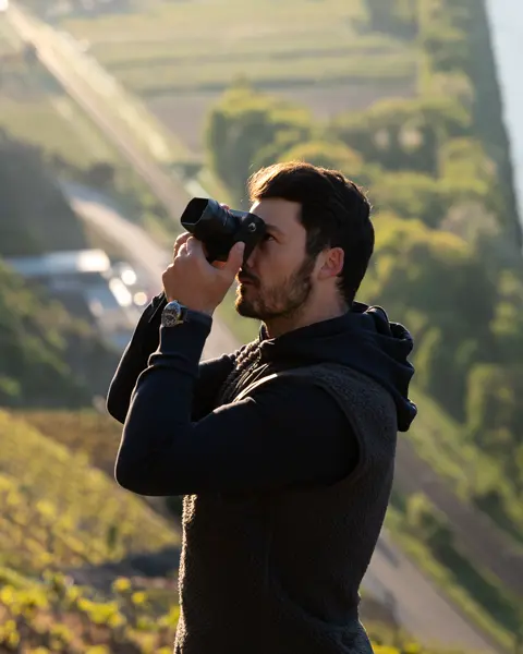 A photographer takes a picture on a sunlit vineyard with a river and trees in the background.