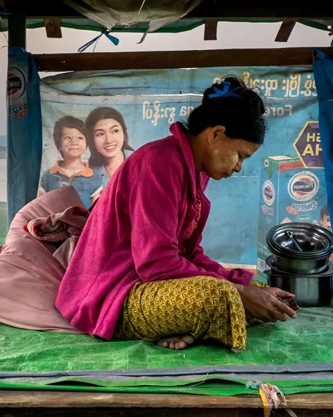 Woman sits on plastic tarp looking at a phone, in background are other people and a river.