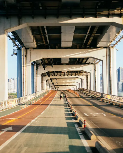 Single cyclist on a bridge with a separate cycle and pedestrian path and road, framed by tall city buildings in the background.