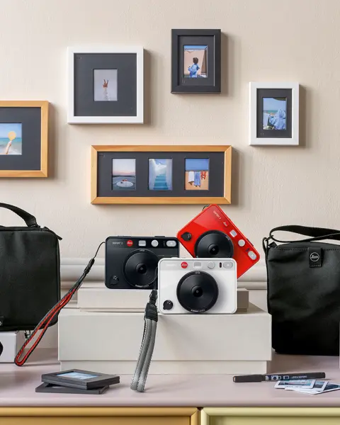 Leica cameras and accessories on a table, with framed photos on the wall in the background.
