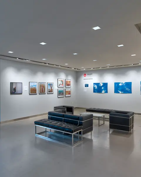 Exhibition room with framed photographs on the wall and black leather armchairs and sofas in the centre of the room.
