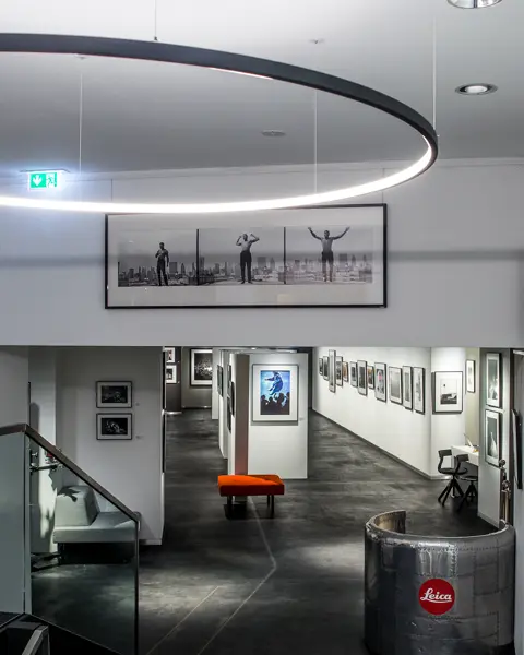 View down a staircase into a long corridor with framed pictures on the wall. At the beginning is a semi-circular metal reception desk with Leica written on it.