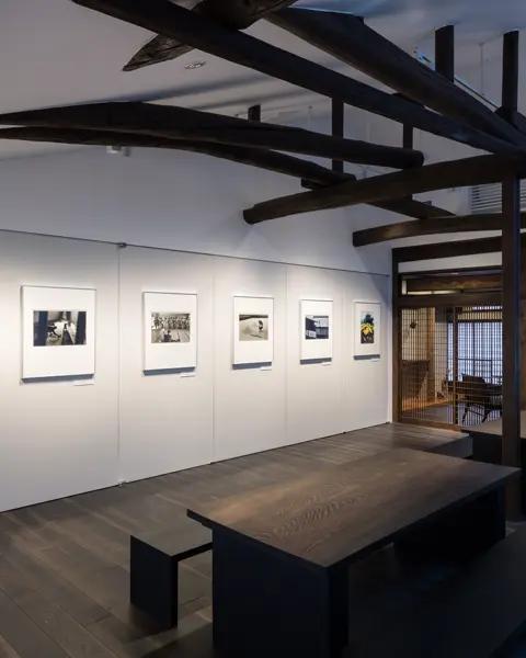 Gallery with black and white photographs, dark wooden beams on the ceiling and a dark wooden seating area. Barred doors lead to other rooms.