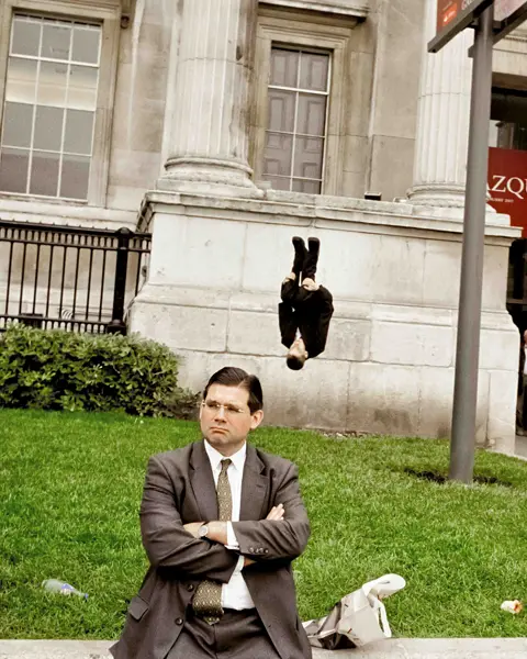 Man in a suit sits on a wall behind him a man in a suit does a somersault