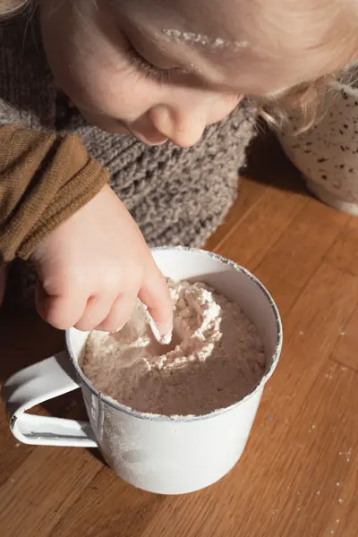 Little girl dips her fingers into a cup full of flour.