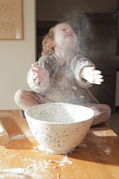 A little girl sits in front of a bowl of flour and claps her hands together with flour.