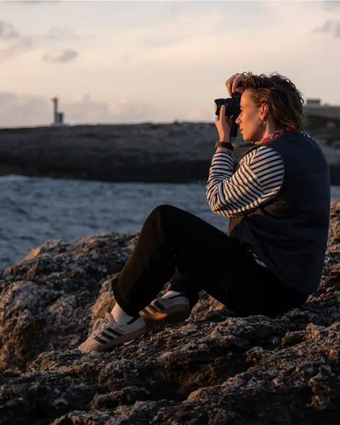 Julia Nimke is sitting on a rock by the sea with her Leica camera, taking photos