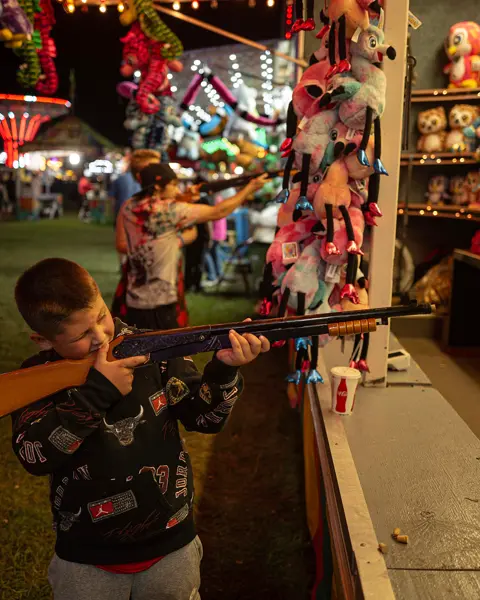 Child aiming at a carnival shooting game, capturing the vibrant atmosphere of a nighttime fairground and family entertainment.