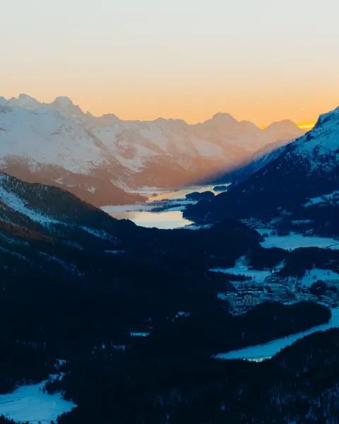 Snow-covered alpine valley at sunrise, with mountain peaks and lakes glowing in the morning light.