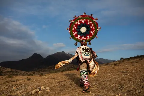 Documentary photograph of a traditional Mexican dancer wearing an elaborate feathered headdress and embroidered costume, walking through a mountainous landscape during a cultural celebration.