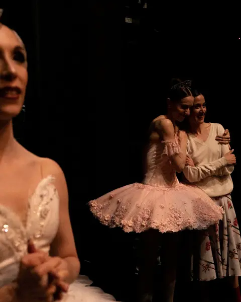 Ballet dancers embracing backstage after a performance while others wait in the wings.