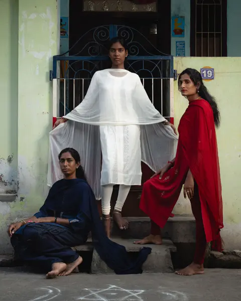 Trois femmes vêtues de bleu, de blanc et de rouge posent devant un bâtiment.