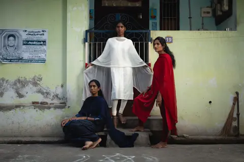 Trois femmes vêtues de bleu, de blanc et de rouge posent devant un bâtiment.