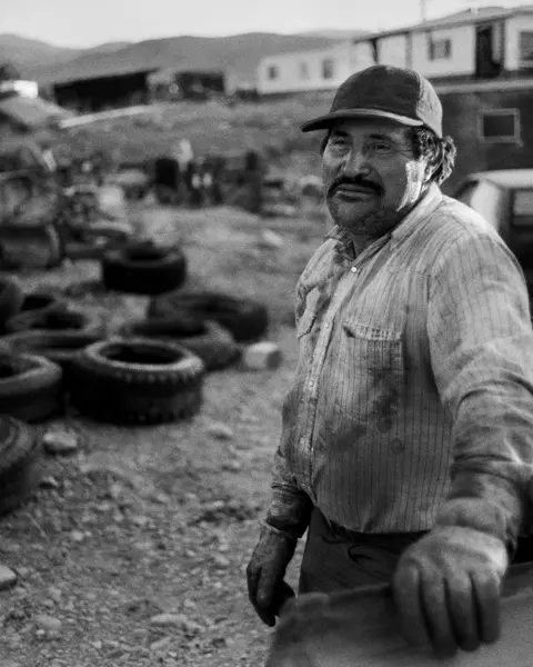 A man in a worn work shirt leans against a pickup truck in a rural scrapyard in Montana. Old tires, machinery, and trailers fill the foreground, with the Rocky Mountains in the distance.