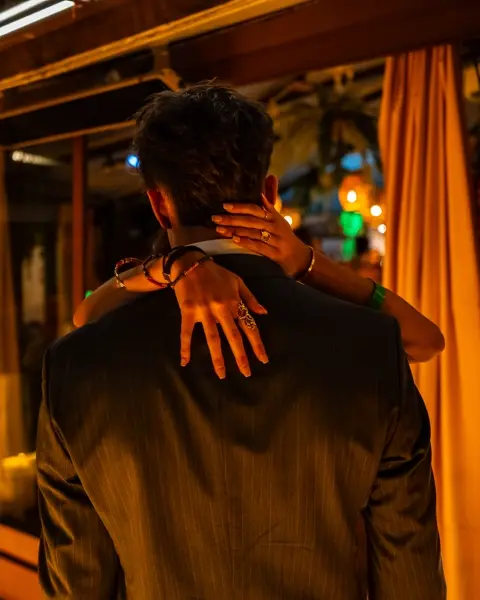 Documentary photograph of a couple embracing at a warmly lit evening celebration, capturing intimacy, human connection, and contemporary social life.
