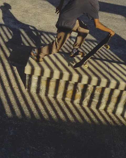 Skateboarder carrying a board through dramatic striped shadows at a skatepark, capturing movement, urban culture and graphic light.