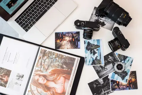 Flat lay of cameras, lenses, prints, and a photography portfolio beside a laptop.