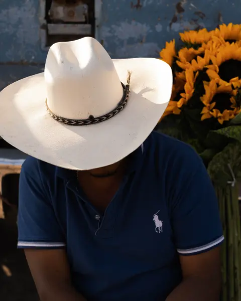 Man in a white cowboy hat sitting beside bright sunflowers and a weathered truck, capturing rural character, color and everyday life.