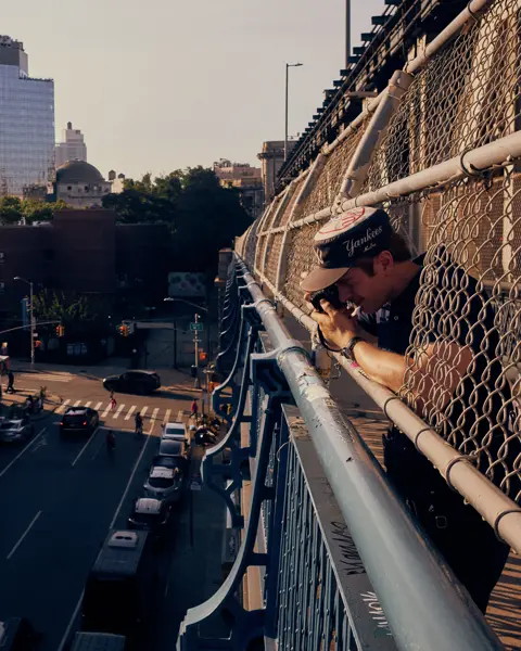 Paul Hepper leaning through the fence of a New York City bridge, taking a street photograph above traffic and urban buildings in warm evening light.