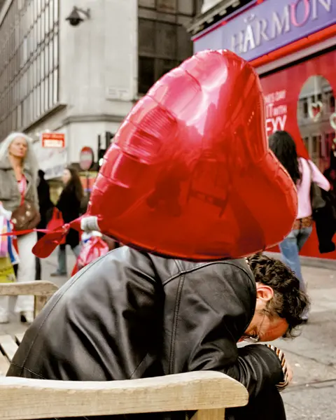 Street scene with a person seated on a bench, partially obscured by a large red heart-shaped balloon, surrounded by pedestrians in an urban setting.