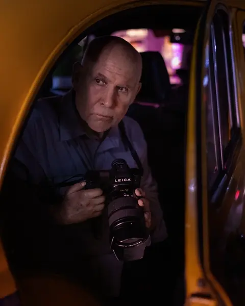 Photographer holding a Leica SL camera while seated inside a yellow taxi at night, illuminated by colorful urban lights.
