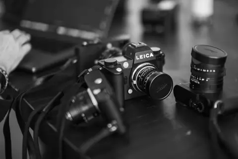 A black-and-white close-up of Leica cameras and lenses arranged on a table, with a Leica SL camera in sharp focus.