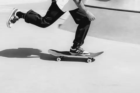 A black-and-white action photograph capturing a skateboarder pushing off on a smooth concrete surface, with dynamic movement and a strong shadow.