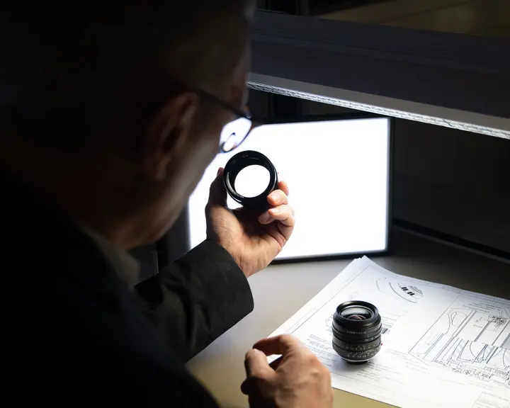 A man inspects a camera lens, technical drawings lie on the table.