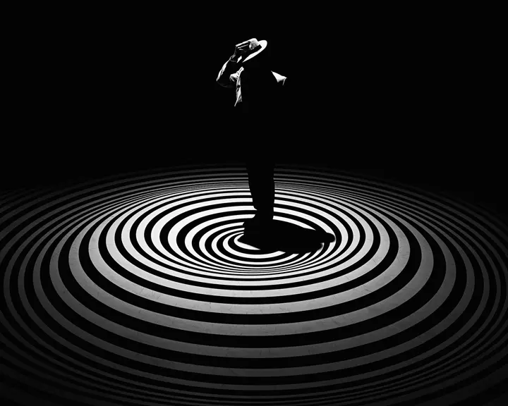 A single person with a hat stands in the centre of concentric circles on a dark background