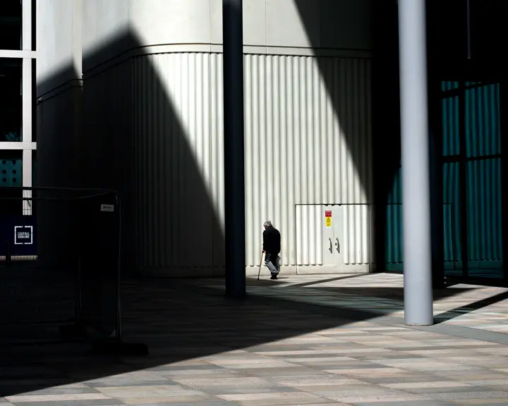 An old man walks in front of a building with shadows beside him.