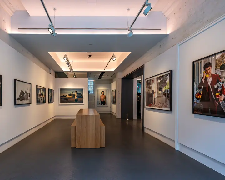 Exhibition room with photographs of various people and a wooden counter and benches in the centre of the room.