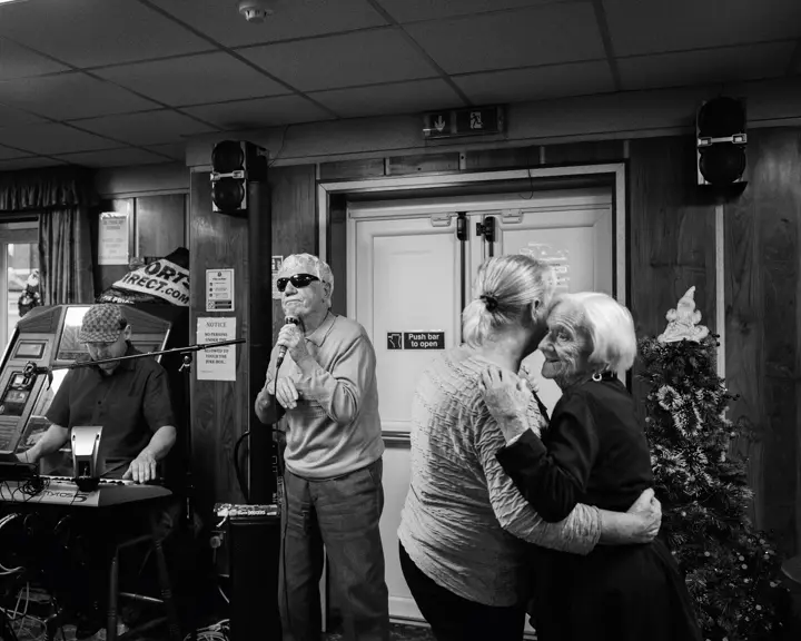 A man sings into a microphone while two older women dance and a man plays the keyboard in the background.