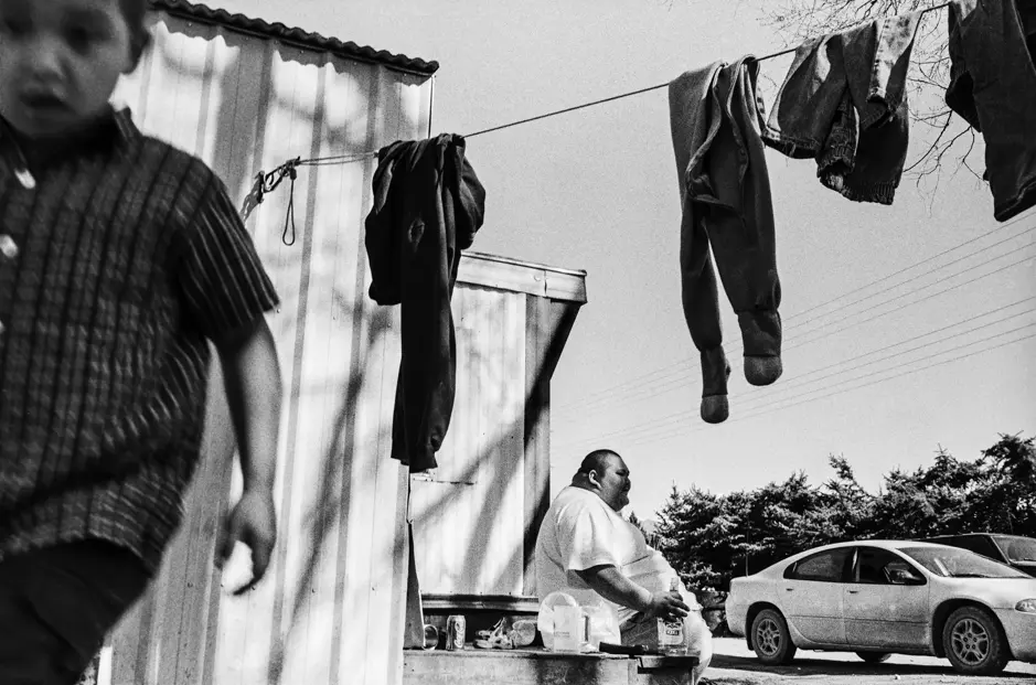 Black-and-white documentary photograph of everyday life in a mobile home community along Highway 93 in Montana, featuring a man outside his trailer, laundry hanging overhead, and a child walking past.