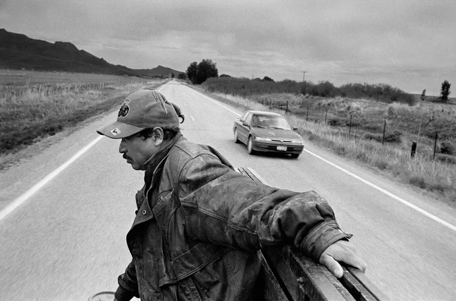 Black-and-white documentary photograph of a man riding in the back of a pickup truck on Highway 93, capturing rural life and open landscapes in Montana, USA.