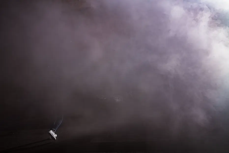 Documentary photograph of a person emerging through dense smoke at night, using dramatic light and atmosphere to evoke mystery, movement, and urban nightlife.