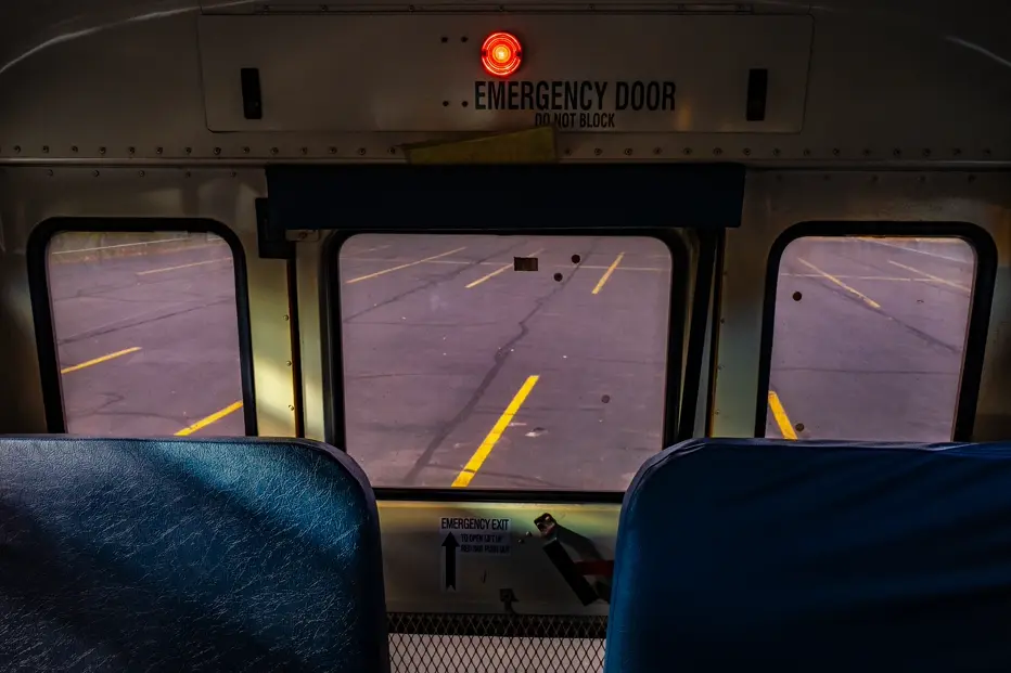 Documentary photograph from inside an empty school bus looking through the emergency exit at an empty parking lot, evoking isolation, travel, and everyday life in the American West.