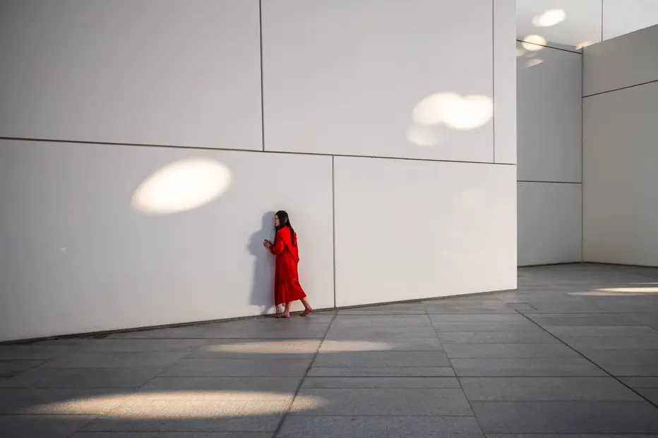 Documentary photograph of a woman in a bright red dress walking barefoot beside a minimalist white wall, creating a striking contrast of color, architecture, and human presence.