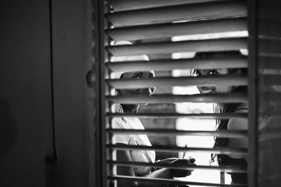 Black-and-white photograph of two women seen through window shutters in Cuba, capturing a candid moment of everyday life and human connection.