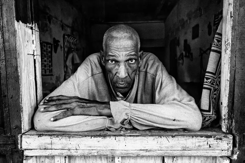 Black-and-white portrait of an older man leaning from a weathered window in Cuba, offering an intimate glimpse into everyday life on the island.