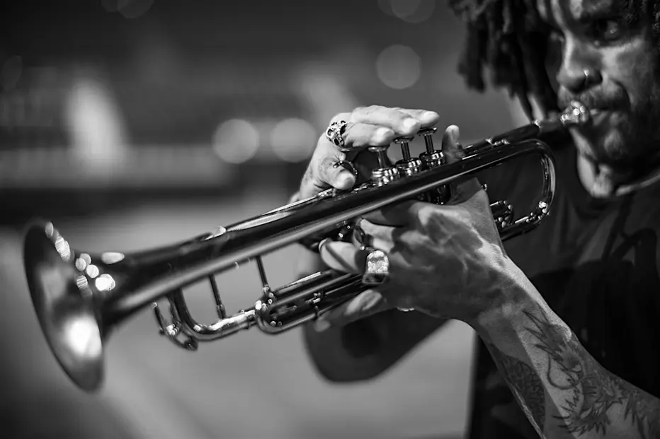 Black-and-white close-up of a trumpet player performing live, highlighting the musician’s hands, tattoos, and instrument.