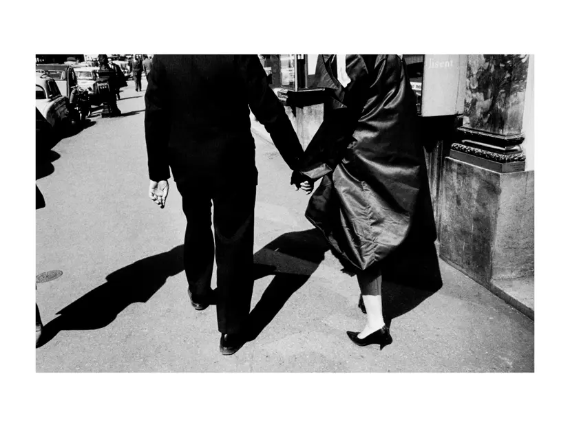 Black-and-white street photograph of a couple walking hand in hand along a sunlit city sidewalk, framed by strong shadows and passing traffic.